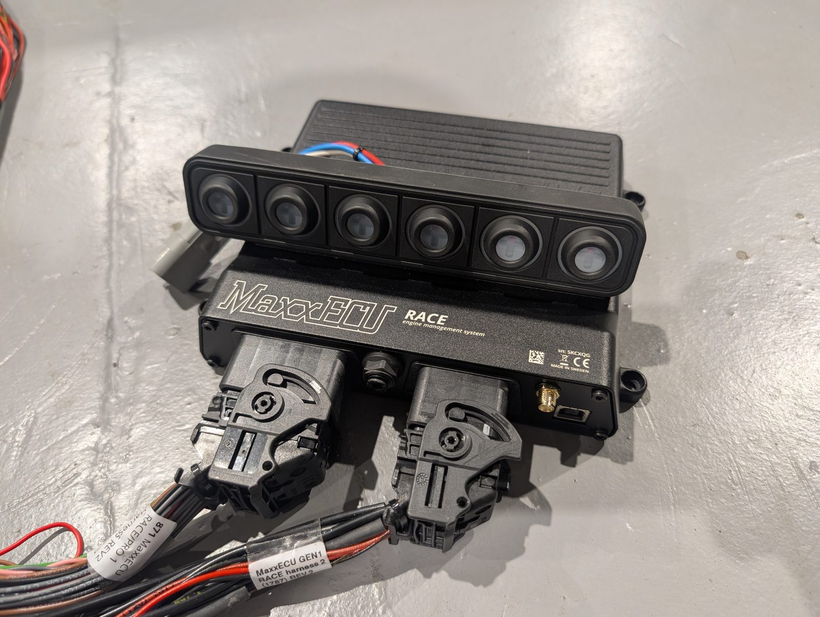 A MaxxECU Race engine control unit with its wiring harness and a six-button switch panel on a workshop floor